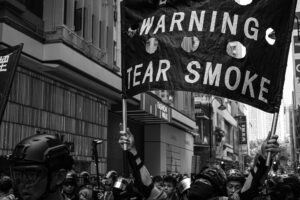 Black and white photo of protestors marching through the streets equipped with eye and head protective gear. One person is holding a banner that warns of tear smoke.